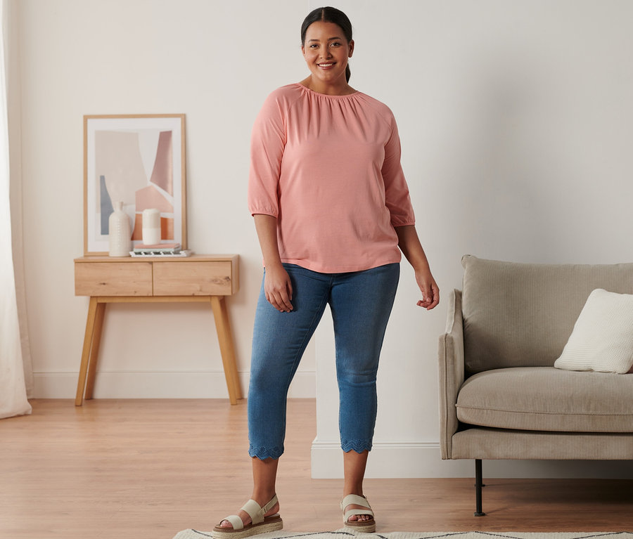 Une femme debout portant une sandale à plateau en cuir, un tregging 7/8 à broderie ajourée et un t-shirt manches 3/4 corail.