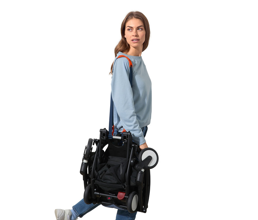 Femme portant une poussette noire pliée sur l'épaule à l'aide d'une bandoulière multifonction.
