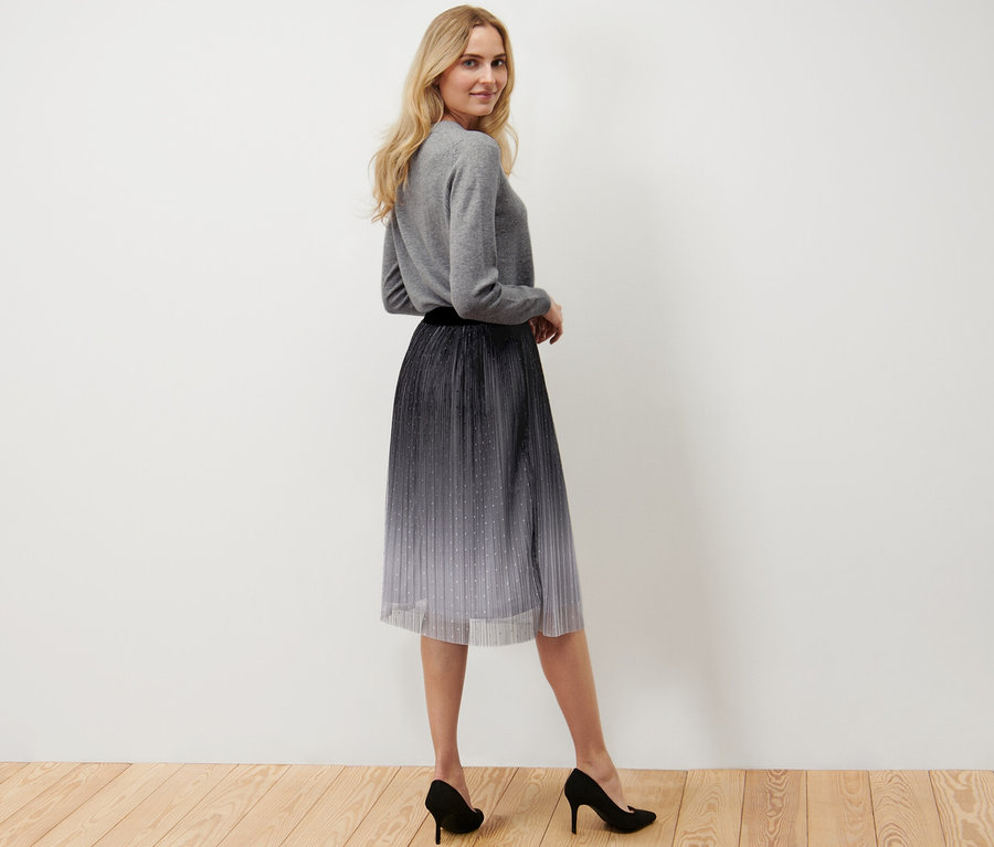Une femme pose et porte une jupe en tulle plissée avec un dégradé, un pull gris et des talons hauts noirs.