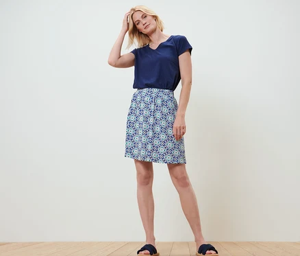 Une femme blonde se tient debout, une main dans les cheveux. Elle porte un t-shirt bleu marine et une jupe en jersey avec un imprimé intégral.