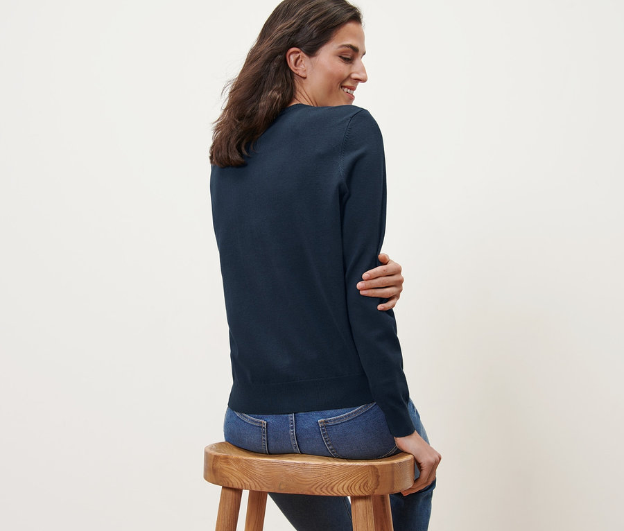 Une femme est assise de côté sur un tabouret en bois et porte un cardigan bleu marine en maille fine et un jean.