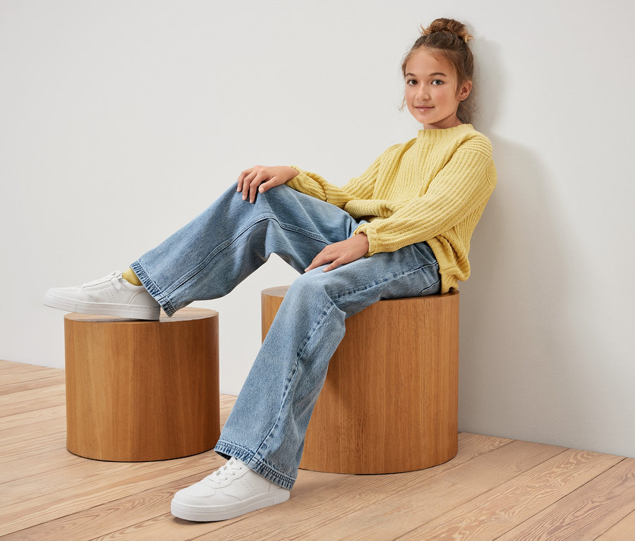 Une jeune fille est assise sur un tabouret en bois, portant un pull jaune et un Jeans pour enfant ‒ Coupe «Lena».