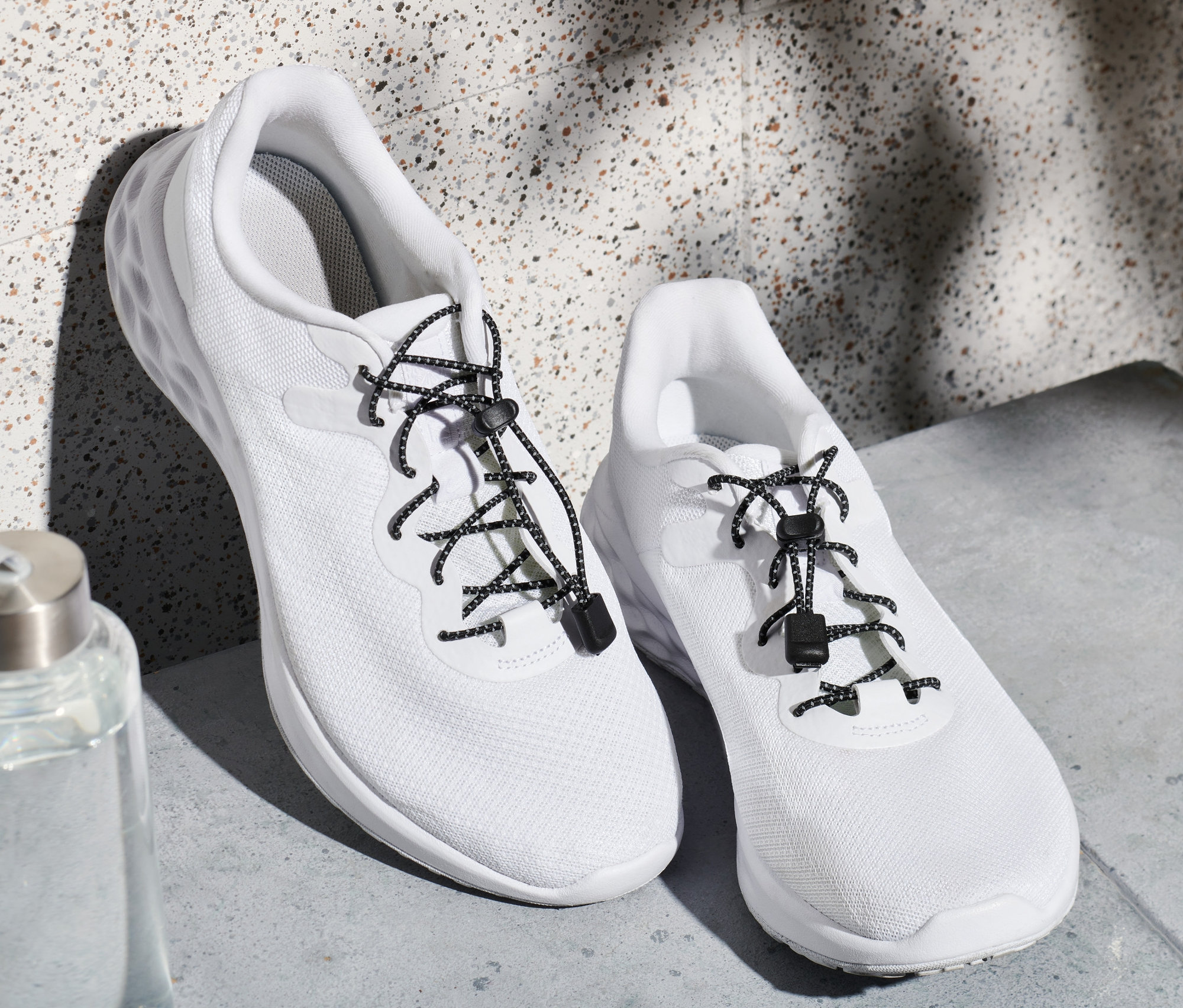 Deux baskets blanches avec des lacets élastiques et une bouteille d'eau.