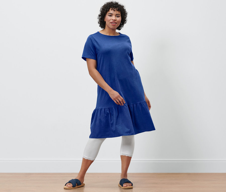 Femme posant en robe en jersey à volants bleu cobalt, legging 3/4 blanc avec bordure en dentelle et sandales confortables.