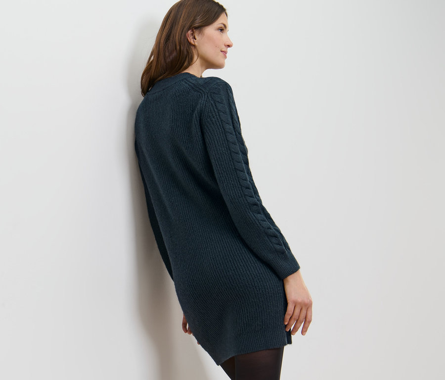 Une femme appuyée contre un mur blanc porte une robe en tricot avec motif torsadé.