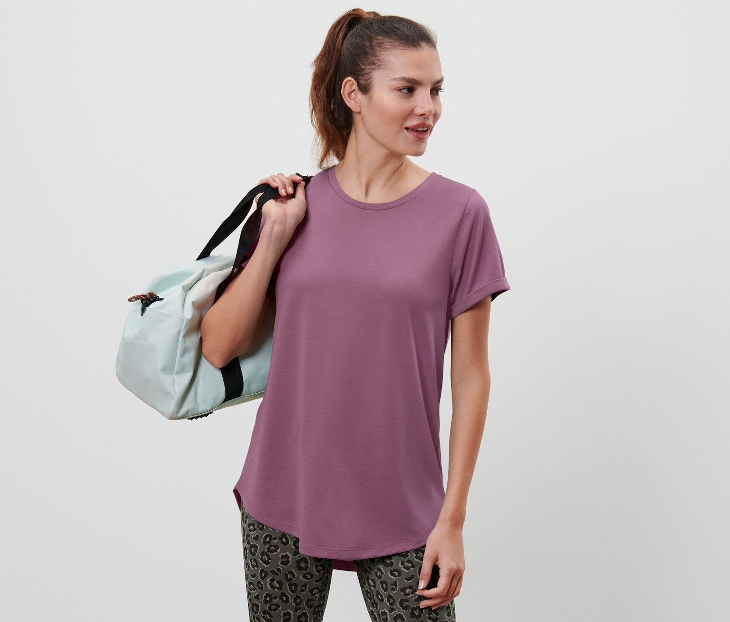 Une femme aux cheveux bruns porte un sac de sport bleu clair sur l'épaule et porte un T-shirt long mauve.