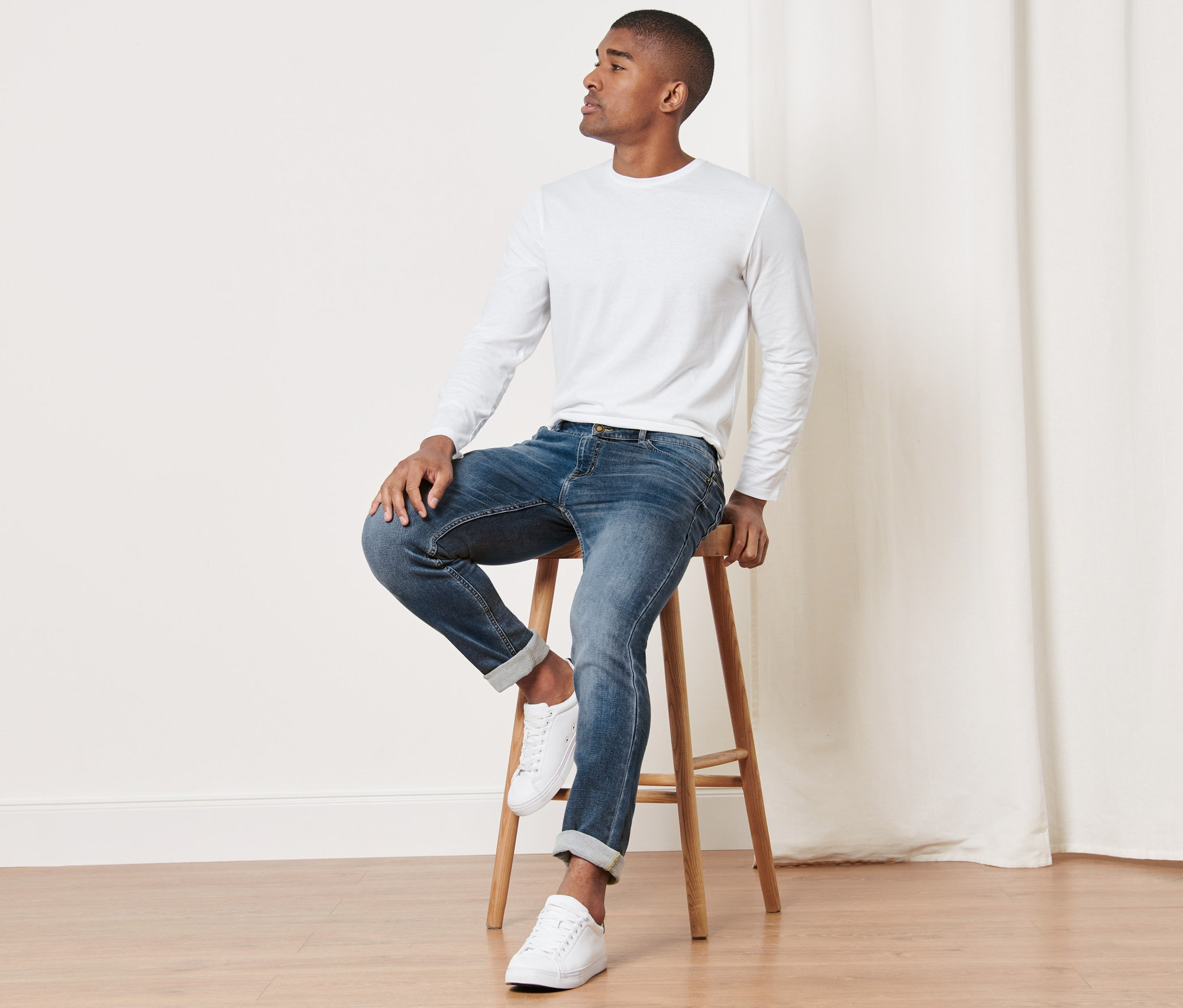 Un homme est assis sur un tabouret en bois et porte un jean, un t-shirt blanc à manches longues et des baskets blanches.