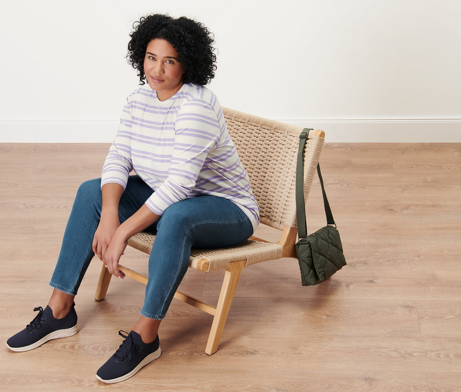 Une femme est assise sur une chaise en osier et porte un jegging, une chemise à manches longues rayée lilas-crème et un sac matelassé à bandoulière.