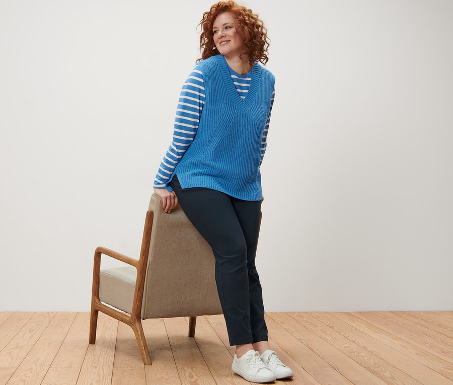 Une femme aux cheveux roux bouclés s'appuie sur une chaise, portant un t-shirt manches longues à rayures, un pull sans manches en maille et un pantalon en bengaline bleu foncé.