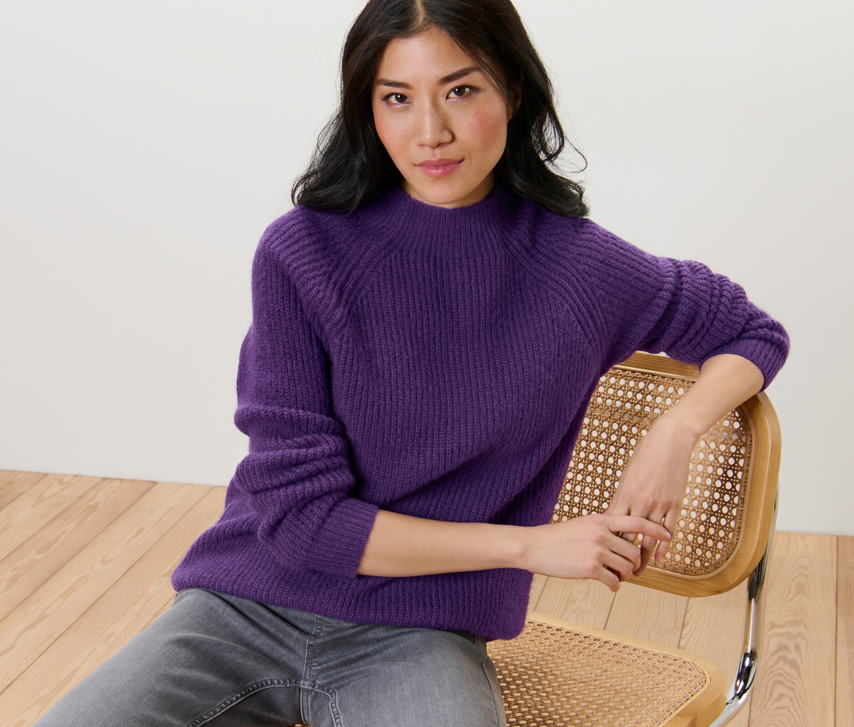 Une femme est assise sur une chaise et porte un pull violet à col droit en grosse maille.