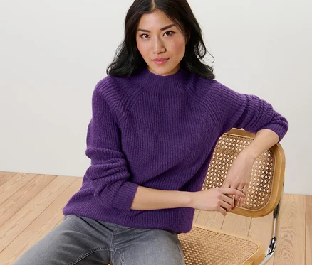 Une femme est assise sur une chaise et porte un pull violet à col droit en grosse maille.