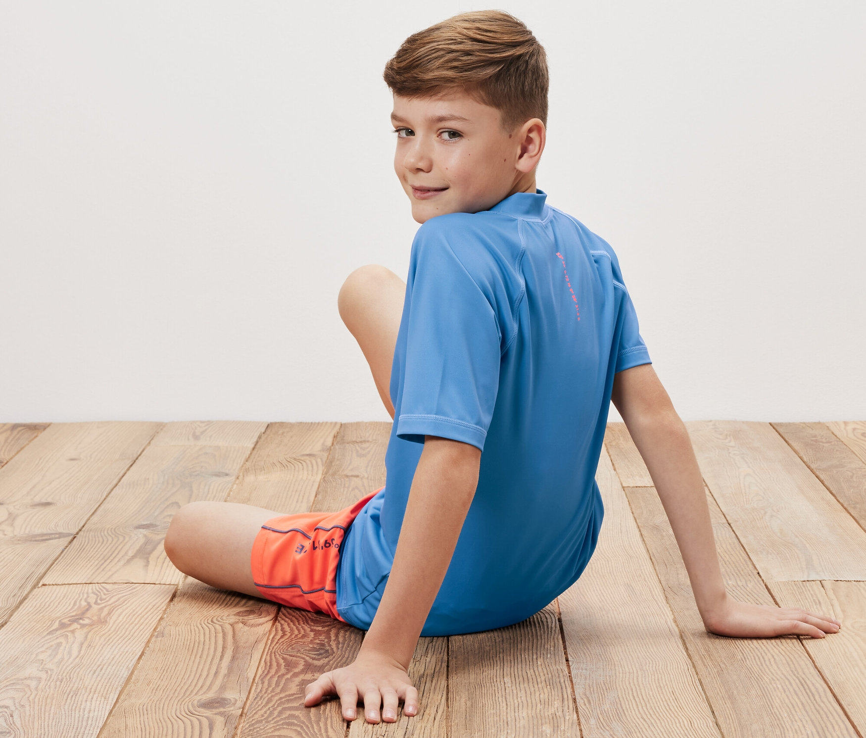 Un garçon assis sur un plancher en bois porte un t-shirt bleu pour enfant avec un indice de protection UV 80 et un short orange.