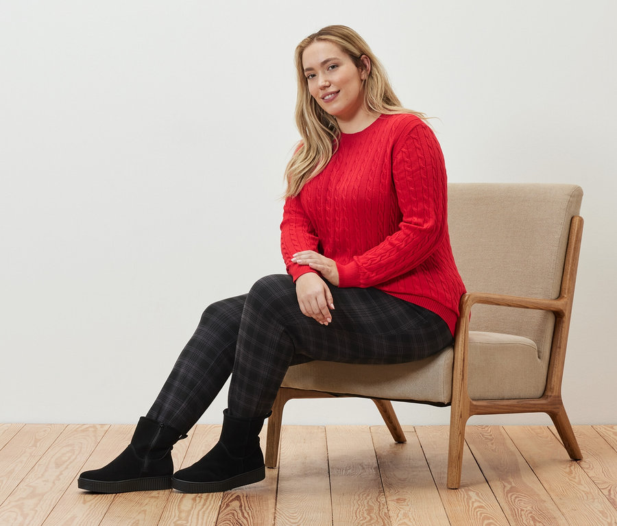 Femme assise sur un fauteuil portant un pull en maille torsadée rouge et un pantalon de jogging à carreaux.