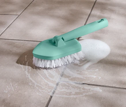 Une brosse turquoise avec des poils blancs est posée sur un carrelage à côté d'un tas de mousse.