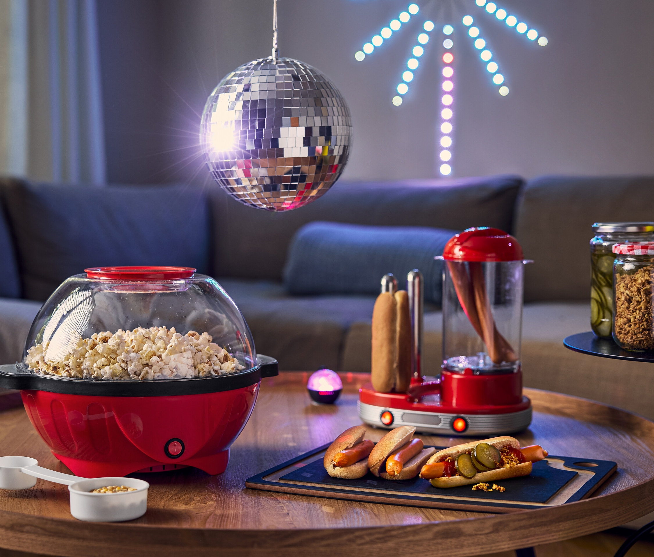 Sur une table, il y a une machine à pop-corn rouge, un cuiseur à hot-dogs et des hot-dogs avec des cornichons, avec une boule disco en arrière-plan.