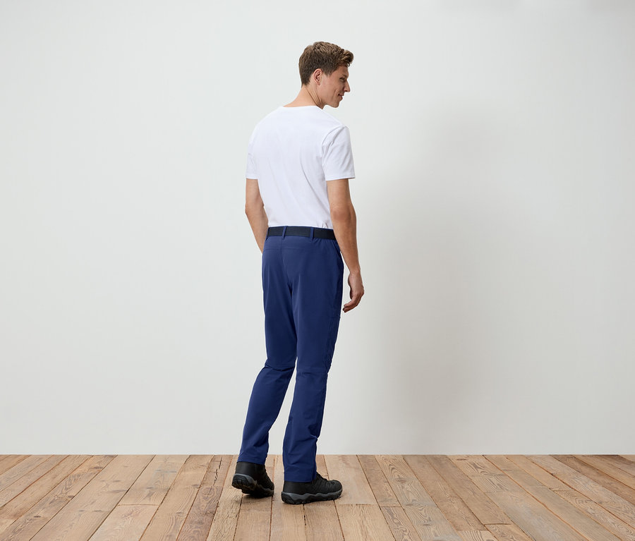 Un homme en t-shirt blanc et pantalon bleu, vu de dos, se tient sur un plancher en bois devant un mur blanc.