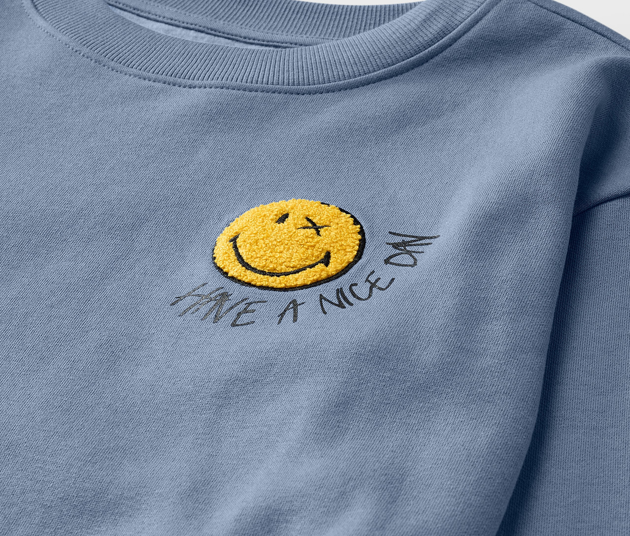 Gros plan sur un sweatshirt pour enfant bleu clair SMILEYWORLD® avec intérieur gratté et smiley jaune.
