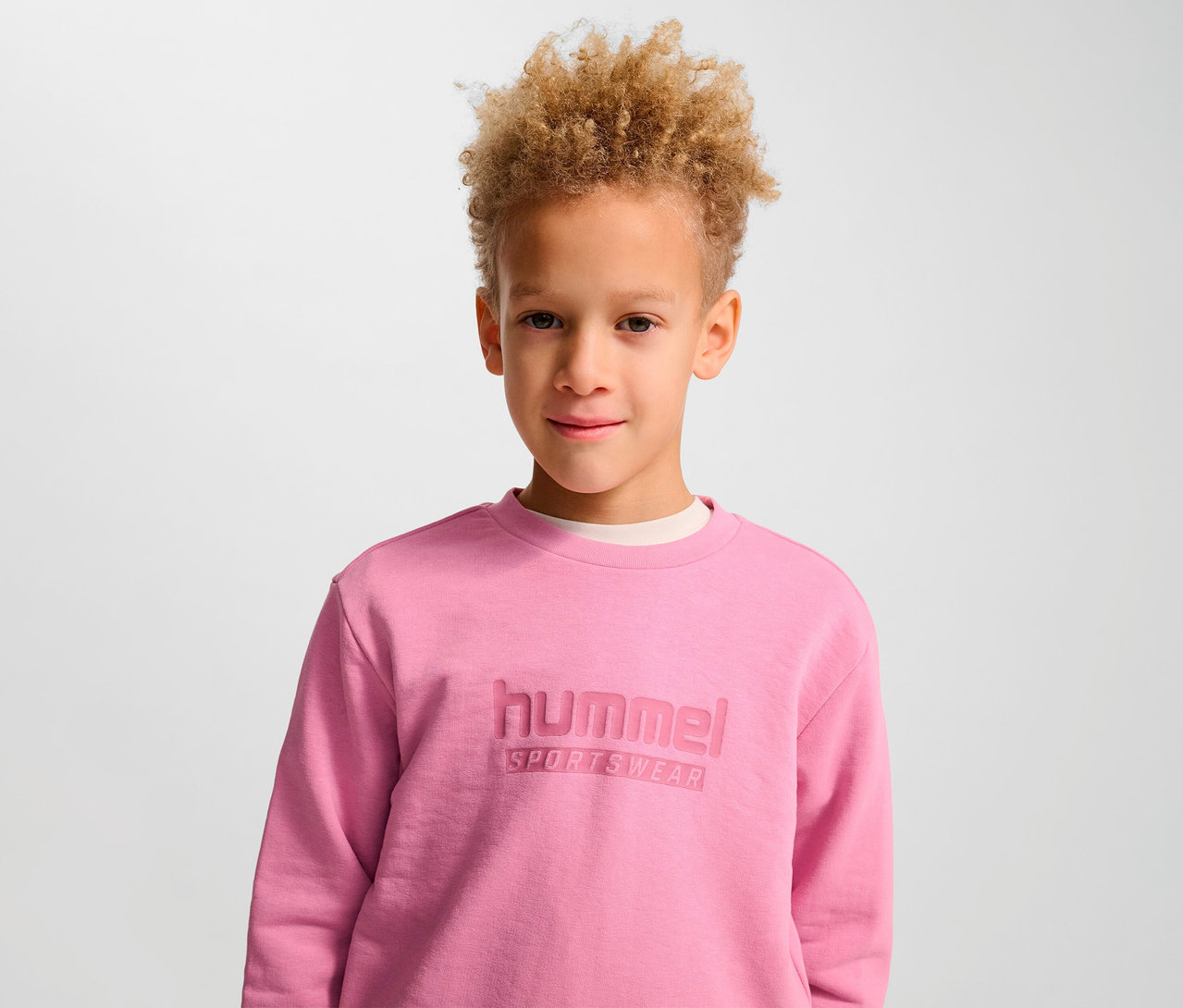 Portrait d'un garçon aux cheveux blonds bouclés portant un sweat-shirt rose HUMMEL HMLJR Base.
