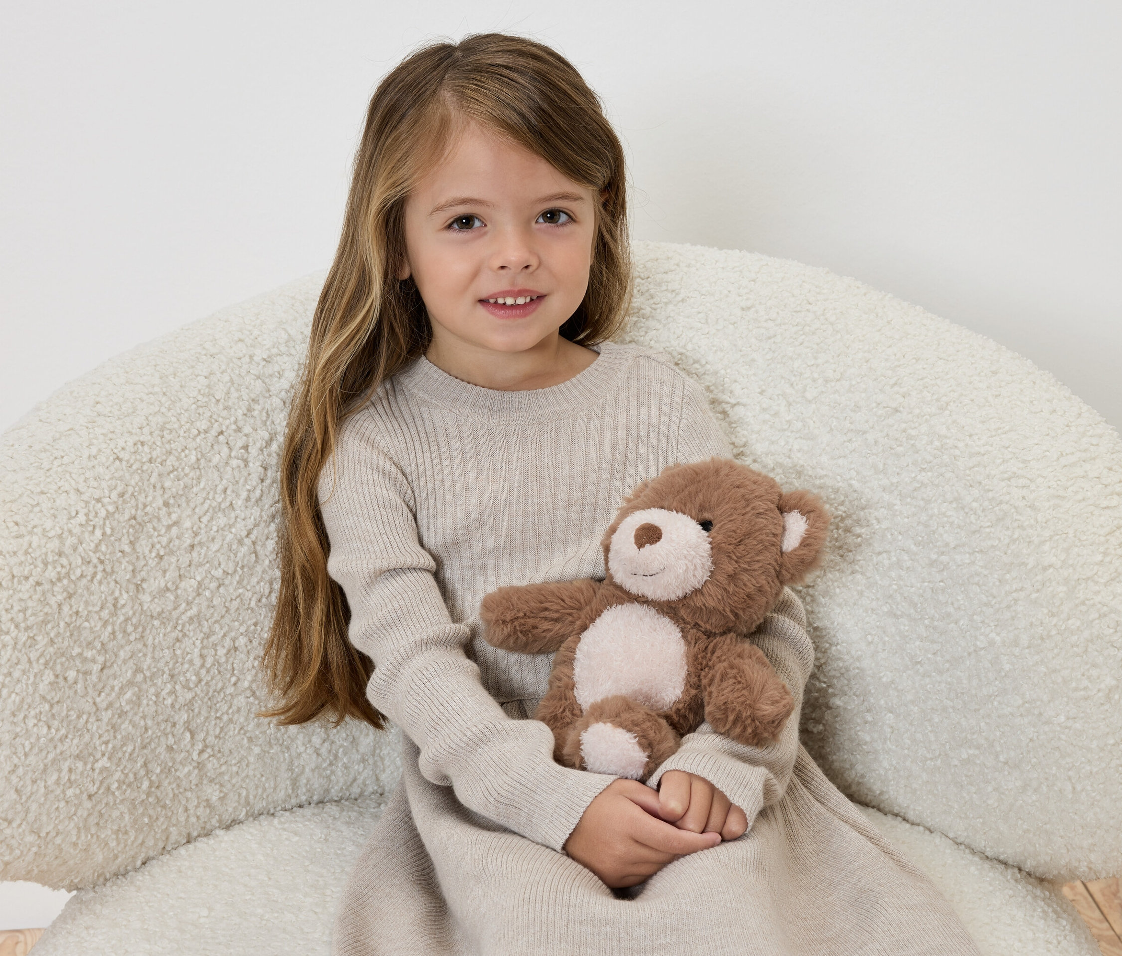 Une fille en robe beige assise sur un fauteuil tenant un ours en peluche marron.