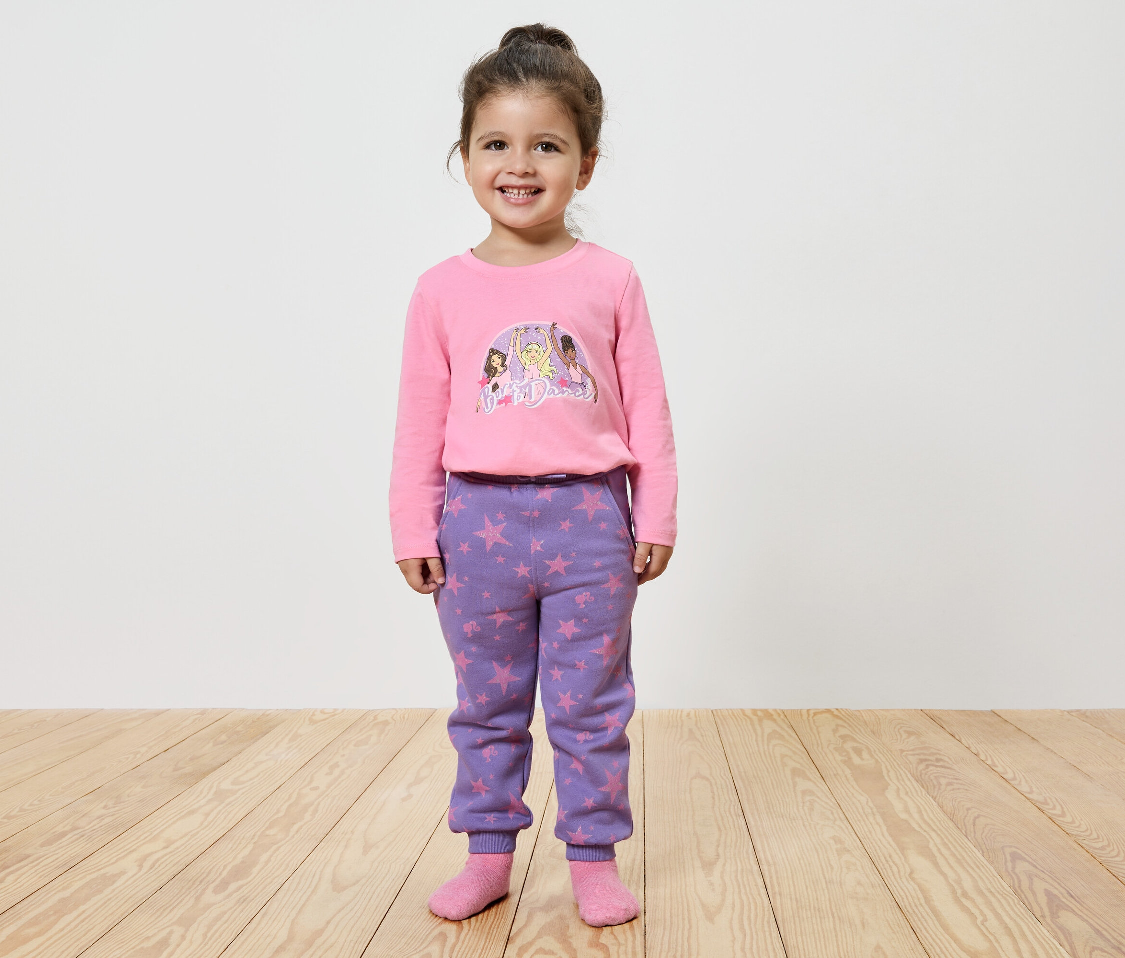 Petite fille souriante posant dans un haut à manches longues rose imprimé et un pantalon violet à étoiles.