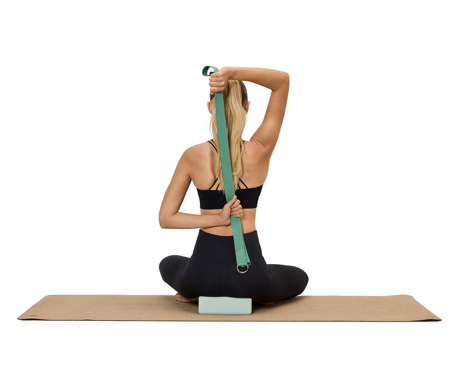 Une femme est assise sur un tapis de yoga avec un bloc en dessous et utilise une sangle de yoga verte pour étirer ses bras derrière son dos.