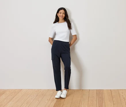 Une femme se tient devant un mur, les mains croisées, portant un t-shirt blanc, un pantalon cargo bleu marine et des baskets blanches.