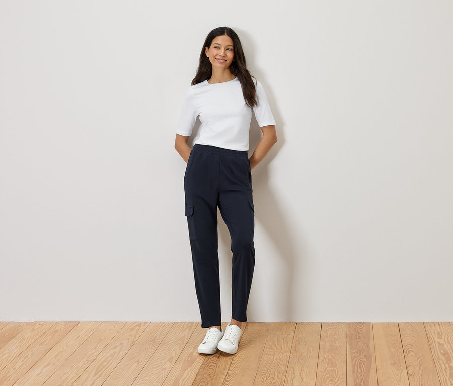 Une femme se tient devant un mur, les mains croisées, portant un t-shirt blanc, un pantalon cargo bleu marine et des baskets blanches.
