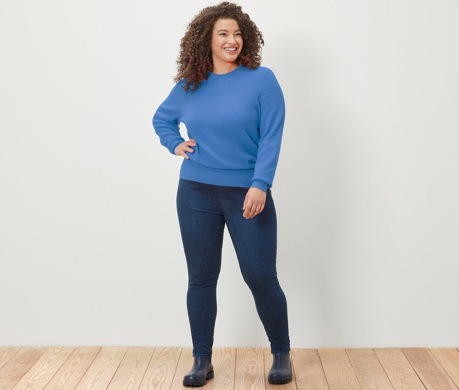 Femme posant sur un plancher en bois avec un pull en maille fine bleu et un jegging.