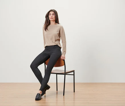 Une femme est assise sur une chaise en bois, portant un pull beige, un pantalon noir et des mocassins noirs.