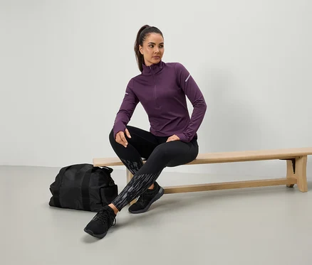 Une femme est assise sur un banc portant un haut de sport violet et un legging noir, un sac de sport noir est posé à côté d'elle.