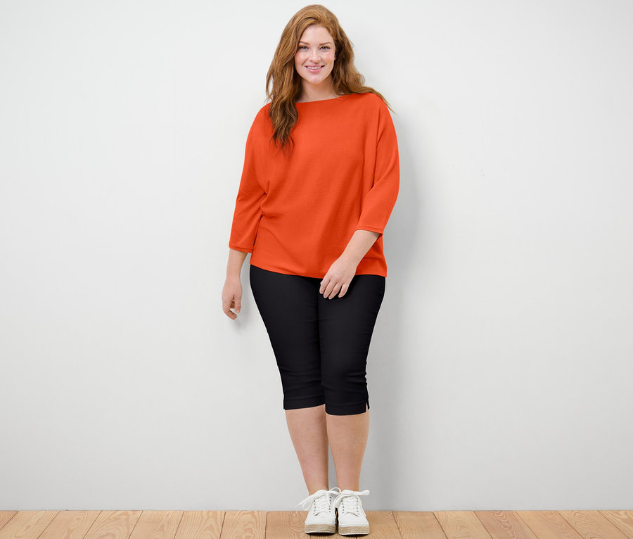 Une femme debout, posant, portant un pull en maille fine orange et un pantalon 3/4 stretch noir.