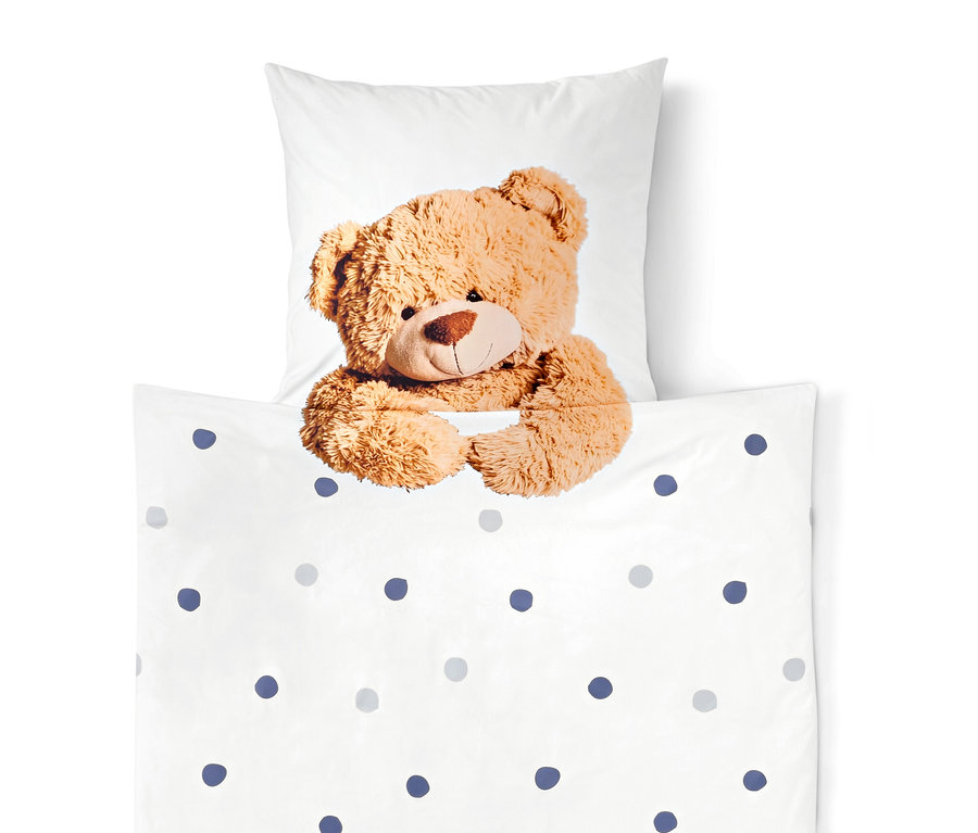 Un ours en peluche marron regarde par-dessus une couverture blanche à pois bleus et gris, avec un oreiller blanc en arrière-plan.