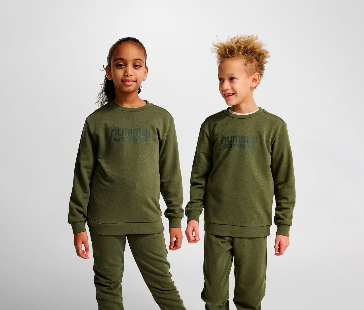 Deux enfants se tiennent côte à côte et portent des sweat-shirts HUMMEL HMLJR Base verts.