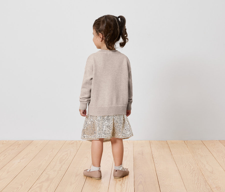 Une petite fille portant un pull beige et une jupe à motifs se tient dos à l'appareil photo.