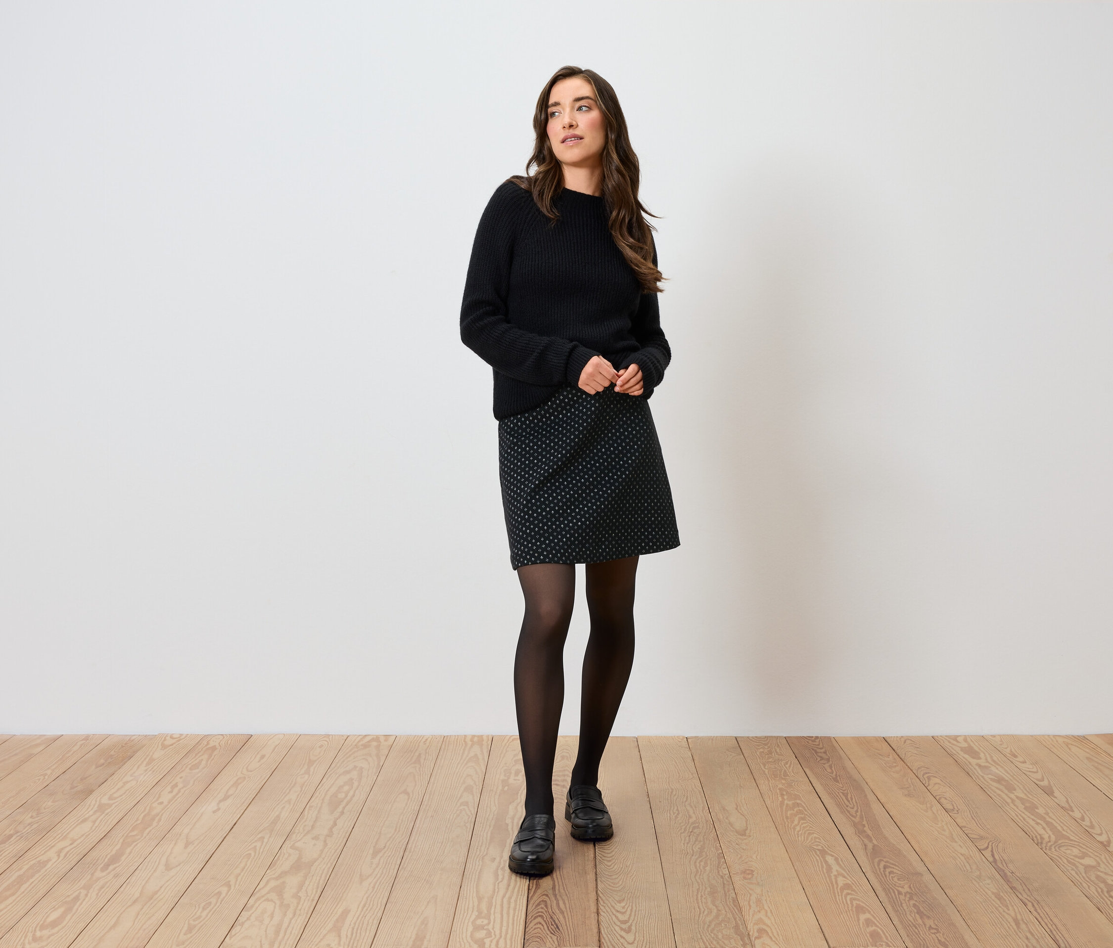 Femme posant en pull noir, jupe à pois, collants noirs et chaussures.