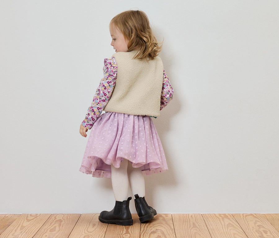 Une petite fille, vue de dos, porte un chemisier à fleurs, un gilet clair, une jupe lilas et des bottines noires.