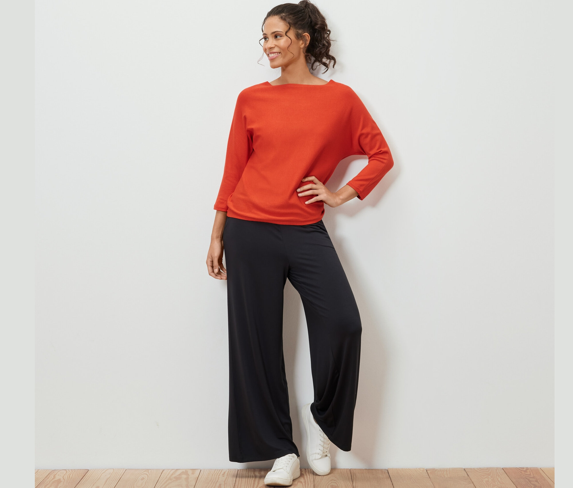 Femme posant en pull en maille fine orange, pantalon noir et baskets blanches.