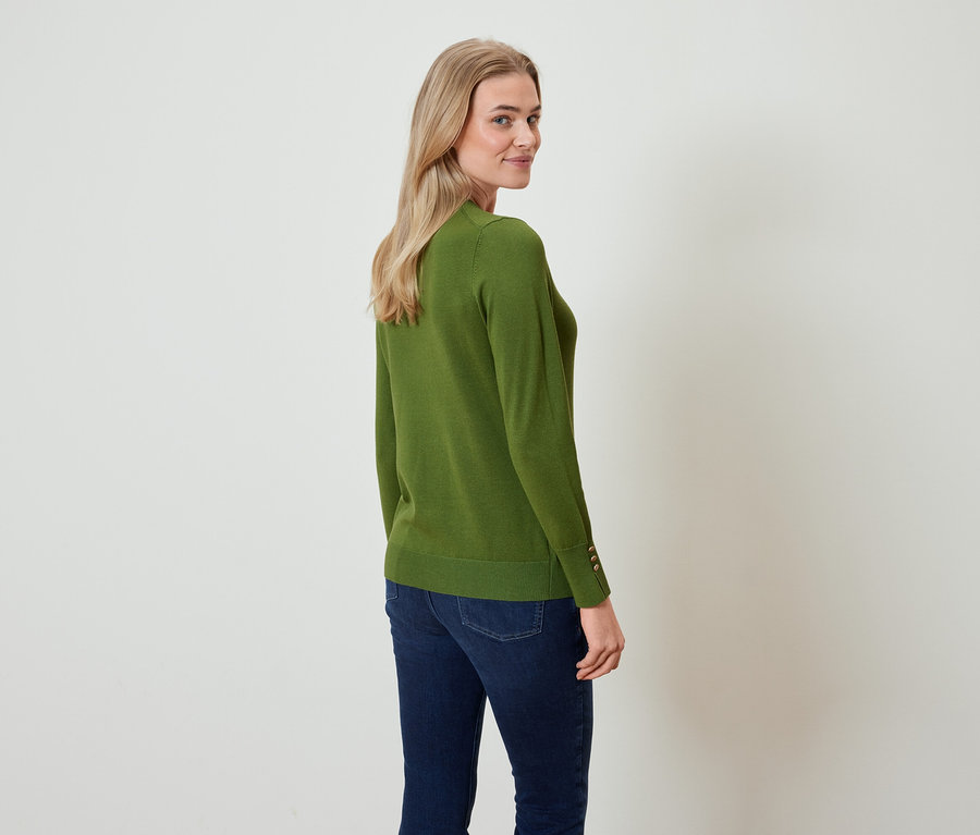 Une femme regarde par-dessus son épaule, portant un pull vert olive et un jean bleu foncé.