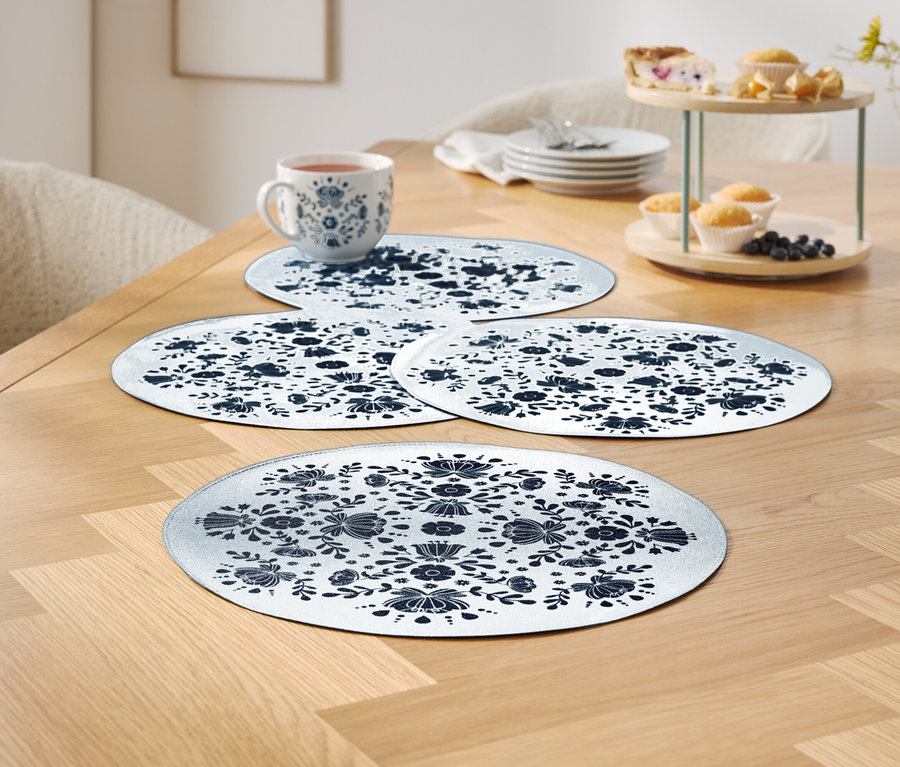 Quatre sets de table blancs à motifs floraux bleus sur une table en bois, une tasse et un plateau de service avec des muffins en arrière-plan.