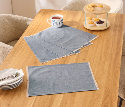 Deux sets de table bleus à bordure blanche sur une table en bois, à côté d'une tasse, d'assiettes et d'un présentoir à gâteaux avec de la nourriture.