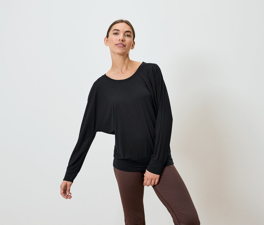 Une femme portant un haut noir à manches longues et un legging marron pose sur un fond blanc.
