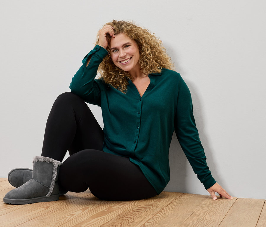 Une femme est assise sur le sol et porte une chemise verte, des leggings noirs et des bottes grises.