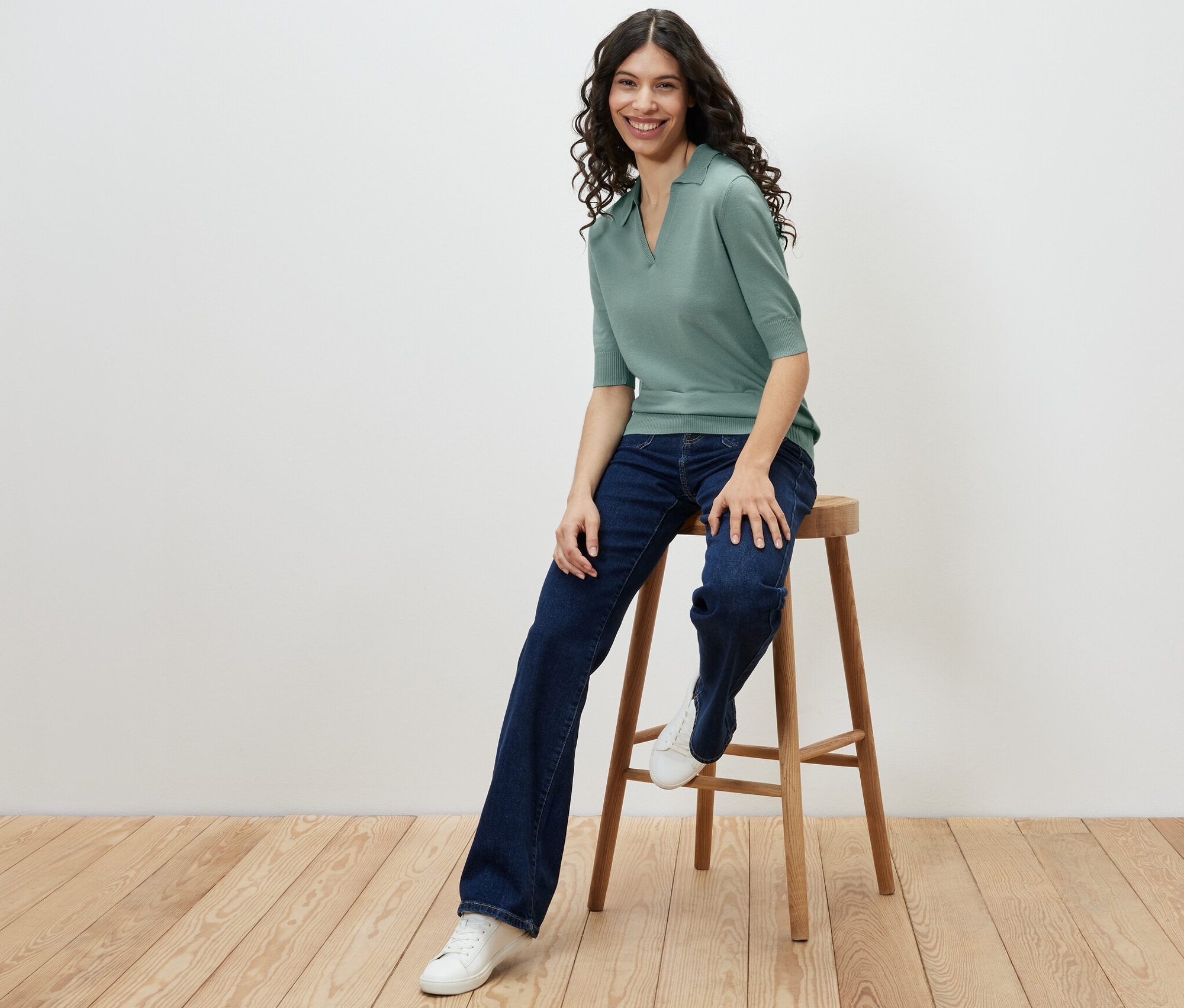 Une femme est assise sur un tabouret et porte un polo en maille fine, un jean et des baskets.