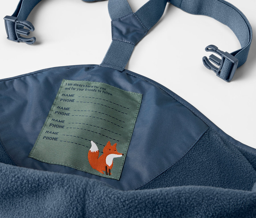 Gros plan sur un pantalon de pluie thermique bleu pour enfant avec bavette et étiquette avec un renard.