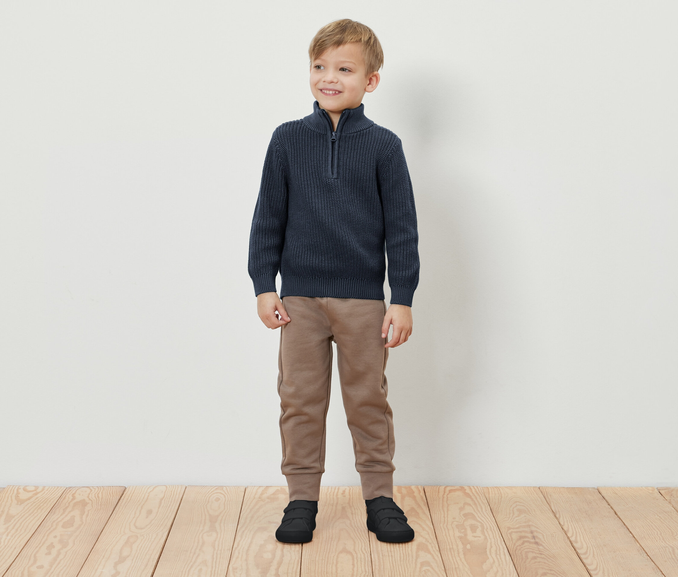 Un garçon se tient debout et sourit, il porte un pull bleu à fermeture éclair, un pantalon de survêtement marron et des chaussures noires.