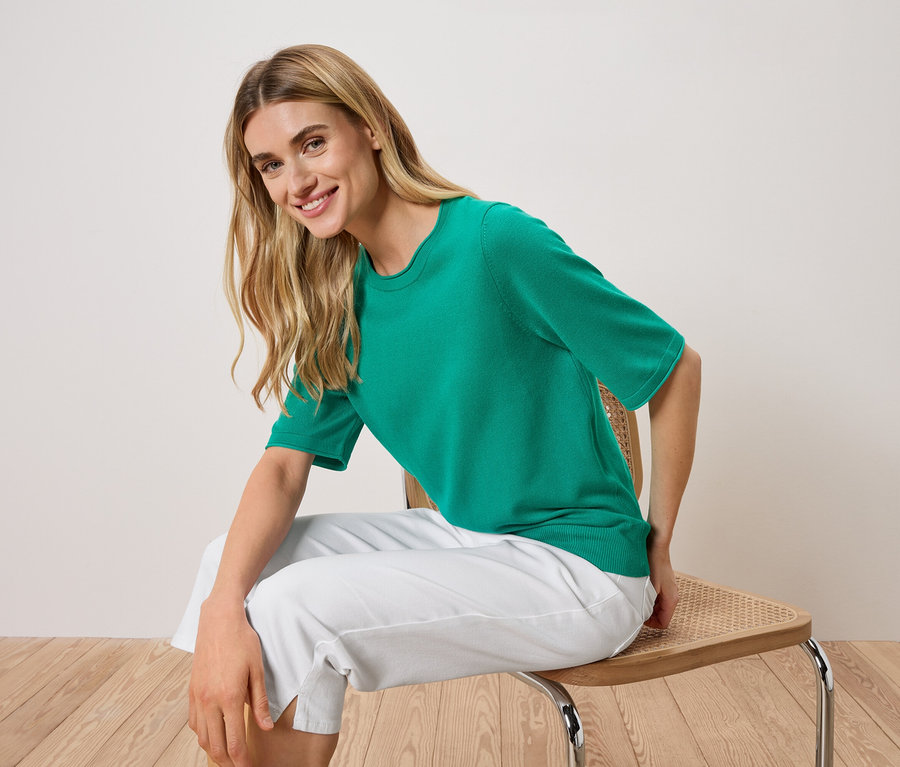 Femme assise sur une chaise, portant un t-shirt vert à manches courtes et un pantalon blanc.