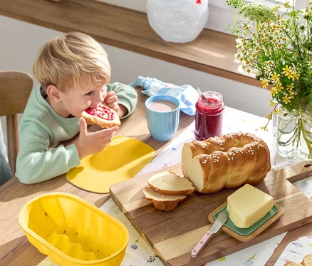 Un garçon blond mange une tartine de confiture à table avec une brioche tressée, du beurre, de la confiture, de la vaisselle et des fleurs.