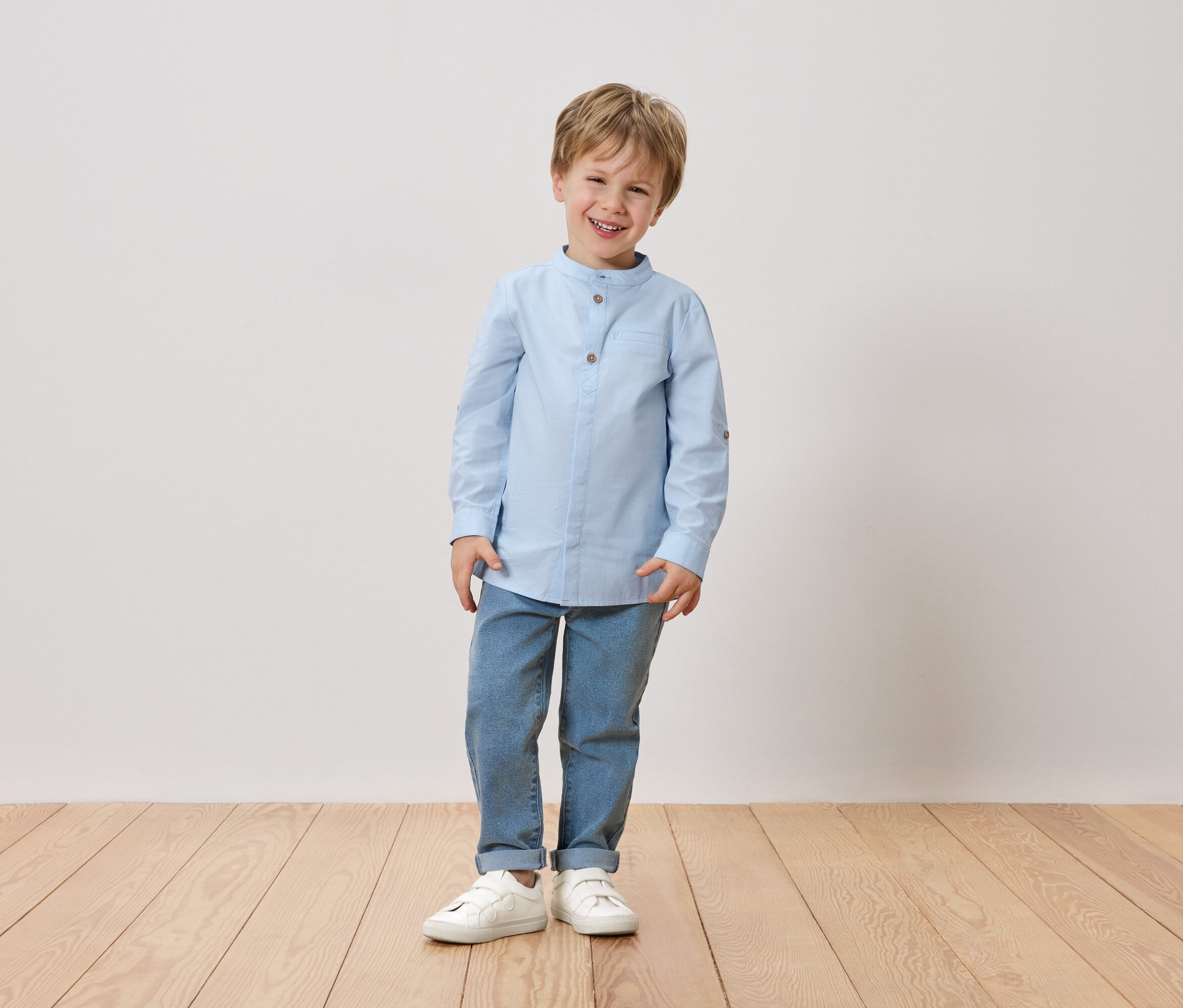Un garçon blond souriant se tient devant un mur blanc. Il porte une chemise bleu clair, un jean et des chaussures blanches.