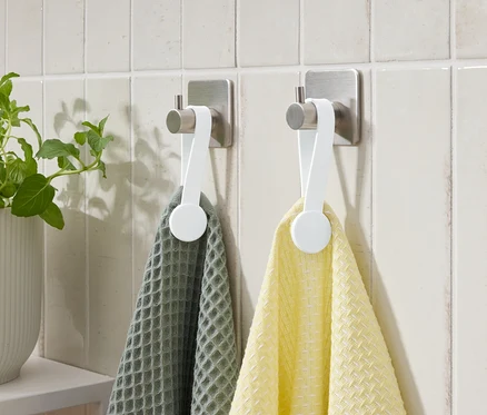 Deux serviettes, une grise et une jaune, sont accrochées à des crochets sur un mur carrelé. Une plante en pot est à gauche.