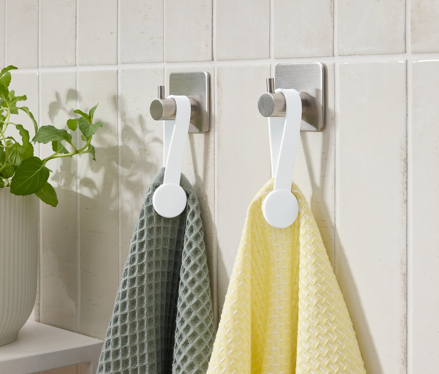 Deux serviettes, une grise et une jaune, sont accrochées à des crochets sur un mur carrelé. Une plante en pot est à gauche.